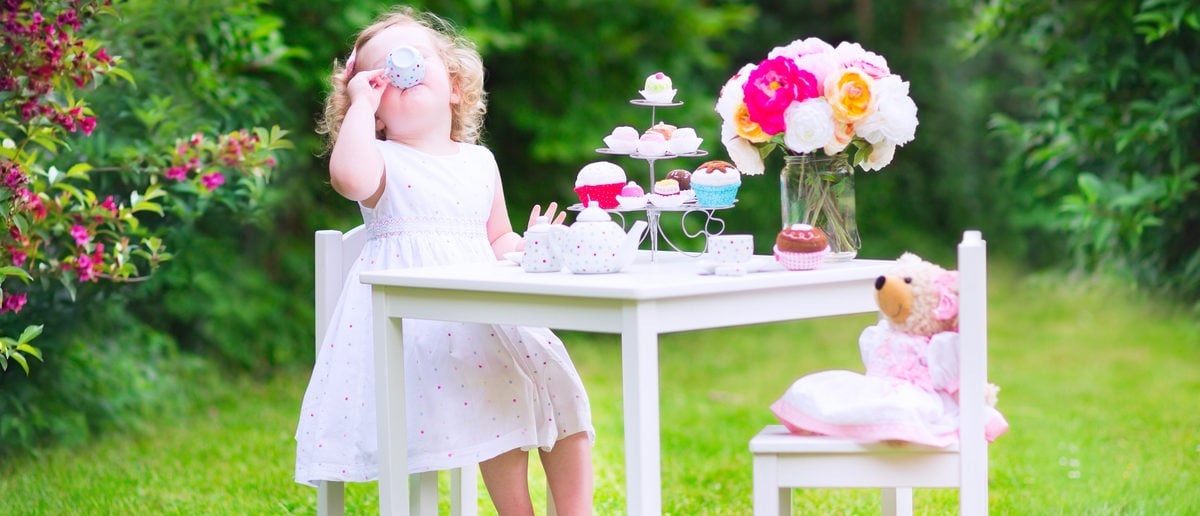 An adorable toddler girl with curly hair is playing tea party with a teddy bear. (Photo: Shutterstock
