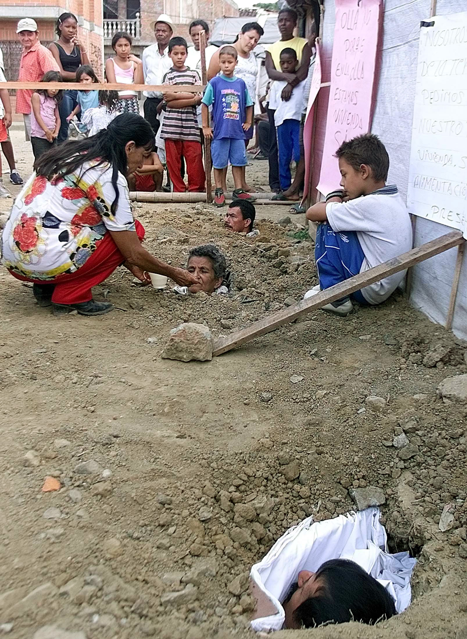 Colombian Maria Gabriela Ruiz, 66, (C) is aided by a woman while she lies buried in the ground up to her neck along with Olmedo Gomez, 42, (bottom) and Nicolas Salazar, 32, during a protest in a popular sector of Cali, July 4, 2003. Three people, two men and a woman, buried themselves three days ago in protest against the government because 150 displaced persons have not been relocated to a safe sector of Cali. REUTERS/Eduardo Munoz