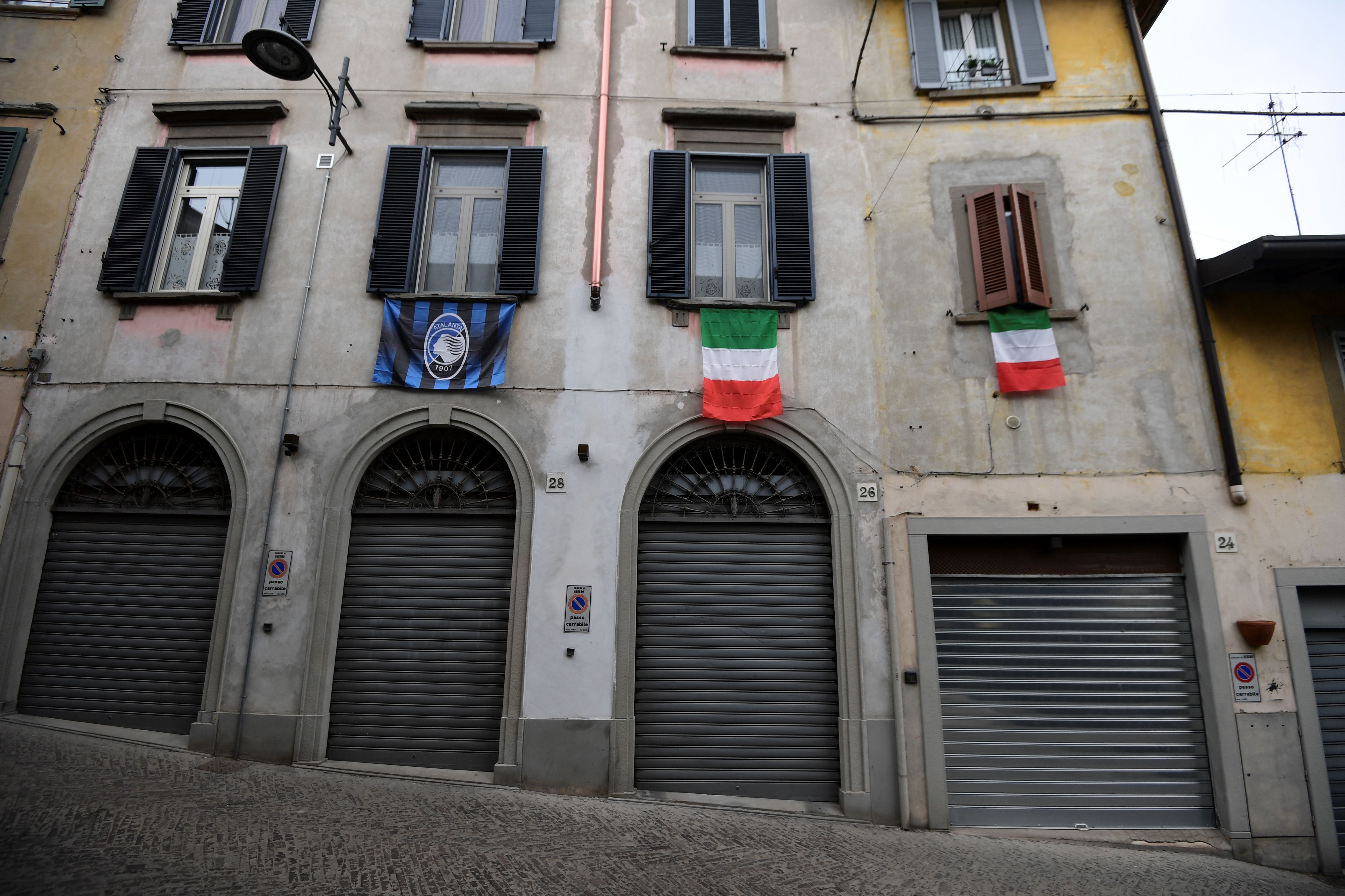 A general view shows Italian flags and an Atalanta Bergamo football team flag (L) hanging over closed shops in a deserted street of Albino near Bergamo, Lombardy, on March 25, 2020, during the country's lockdown following the COVID-19 new coronavirus pandemic. (Photo by MIGUEL MEDINA/AFP via Getty Images)