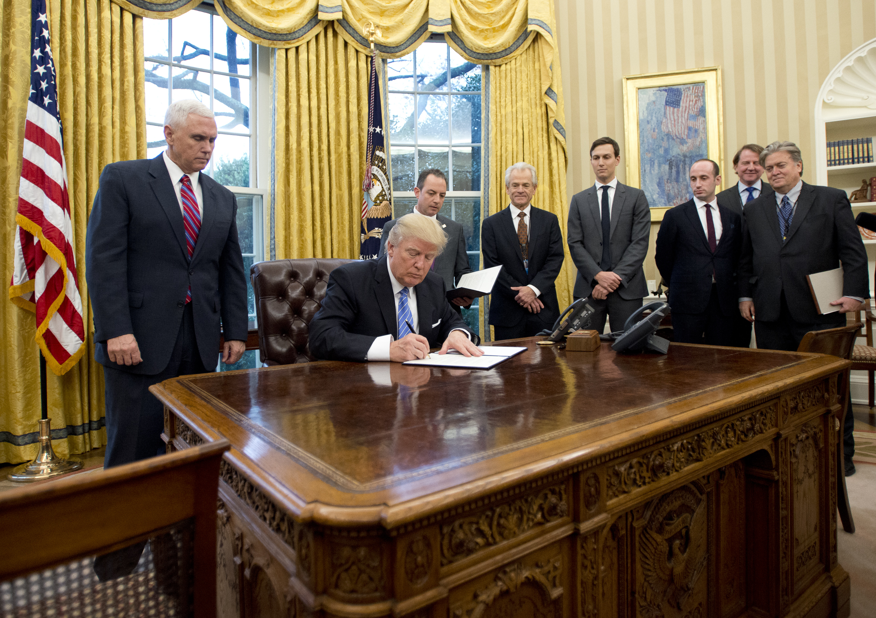 WASHINGTON, DC - JANUARY 23: (AFP OUT) U.S. President Donald Trump signs the first of three Executive Orders in the Oval Office of the White House in Washington, DC on Monday, January 23, 2017. They concerned the withdrawal of the United States from the Trans-Pacific Partnership (TPP), a US Government hiring freeze for all departments but the military, and "Mexico City" which bans federal funding of abortions overseas. Standing behind the President, from left to right: US Vice President Mike Pence; White House Chief of Staff Reince Preibus; Peter Navarro, Director of the National Trade Council; Jared Kushner, Senior Advisor to the President; Steven Miller, Senior Advisor to the President; unknown; and Steve Bannon, White House Chief Strategist. (Photo by Ron Sachs - Pool/Getty Images)