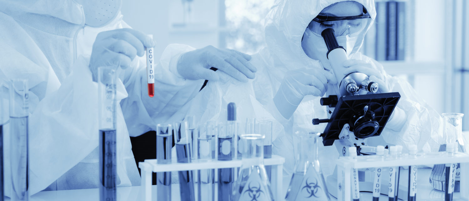 scientist in biohazard protection clothing analyzing covid 19 sample with microscope and holding coronavirus covid 19 blood sample tube on hand in laboratory, coronavirus covid 19 vaccine research (Shutterstock/Mongkolchon Akesin)
