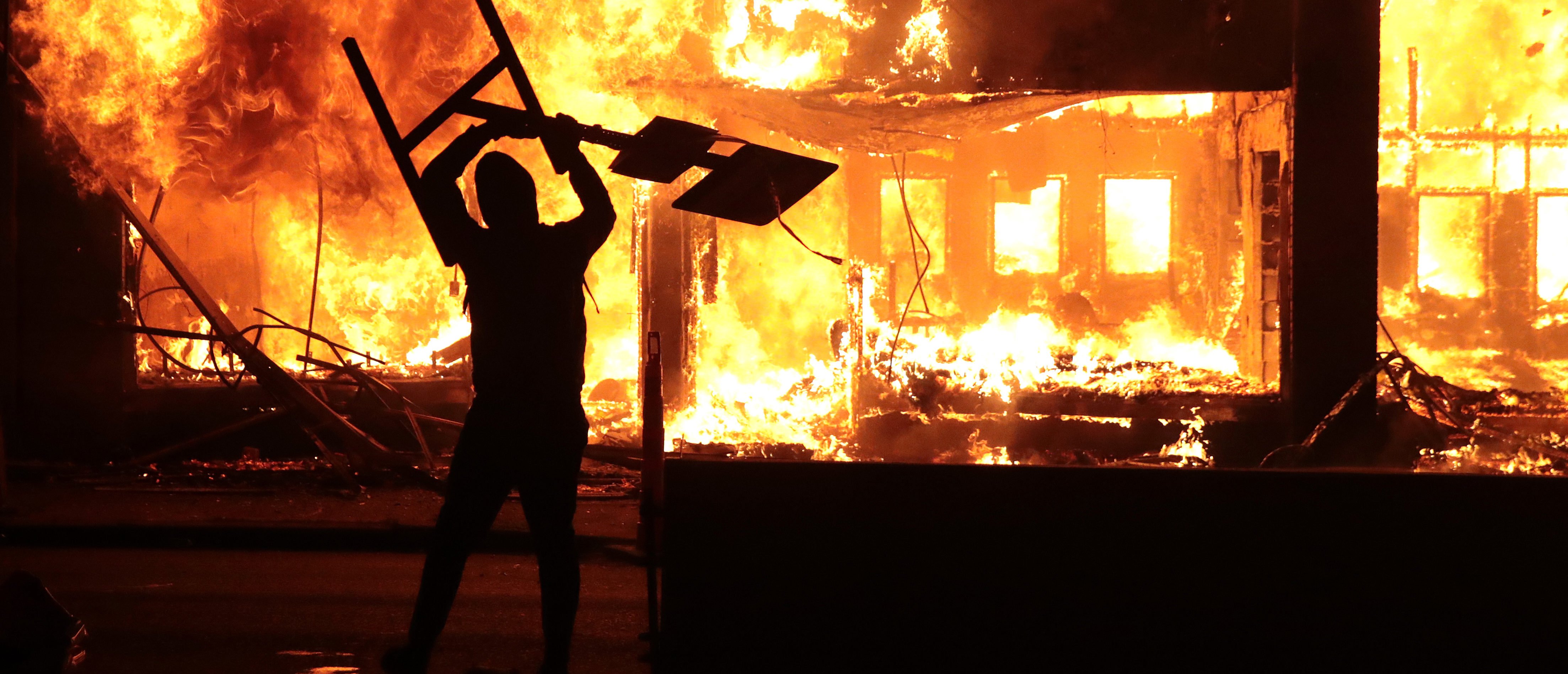 MINNEAPOLIS, MINNESOTA - MAY 29: A man holds up a sign near a burning building during protests sparked by the death of George Floyd while in police custody on May 29, 2020 in Minneapolis, Minnesota. Earlier today, former Minneapolis police officer Derek Chauvin was taken into custody for Floyd's death. Chauvin has been accused of kneeling on Floyd's neck as he pleaded with him about not being able to breathe. Floyd was pronounced dead a short while later. Chauvin and 3 other officers, who were involved in the arrest, were fired from the police department after a video of the arrest was circulated. (Photo by Scott Olson/Getty Images)