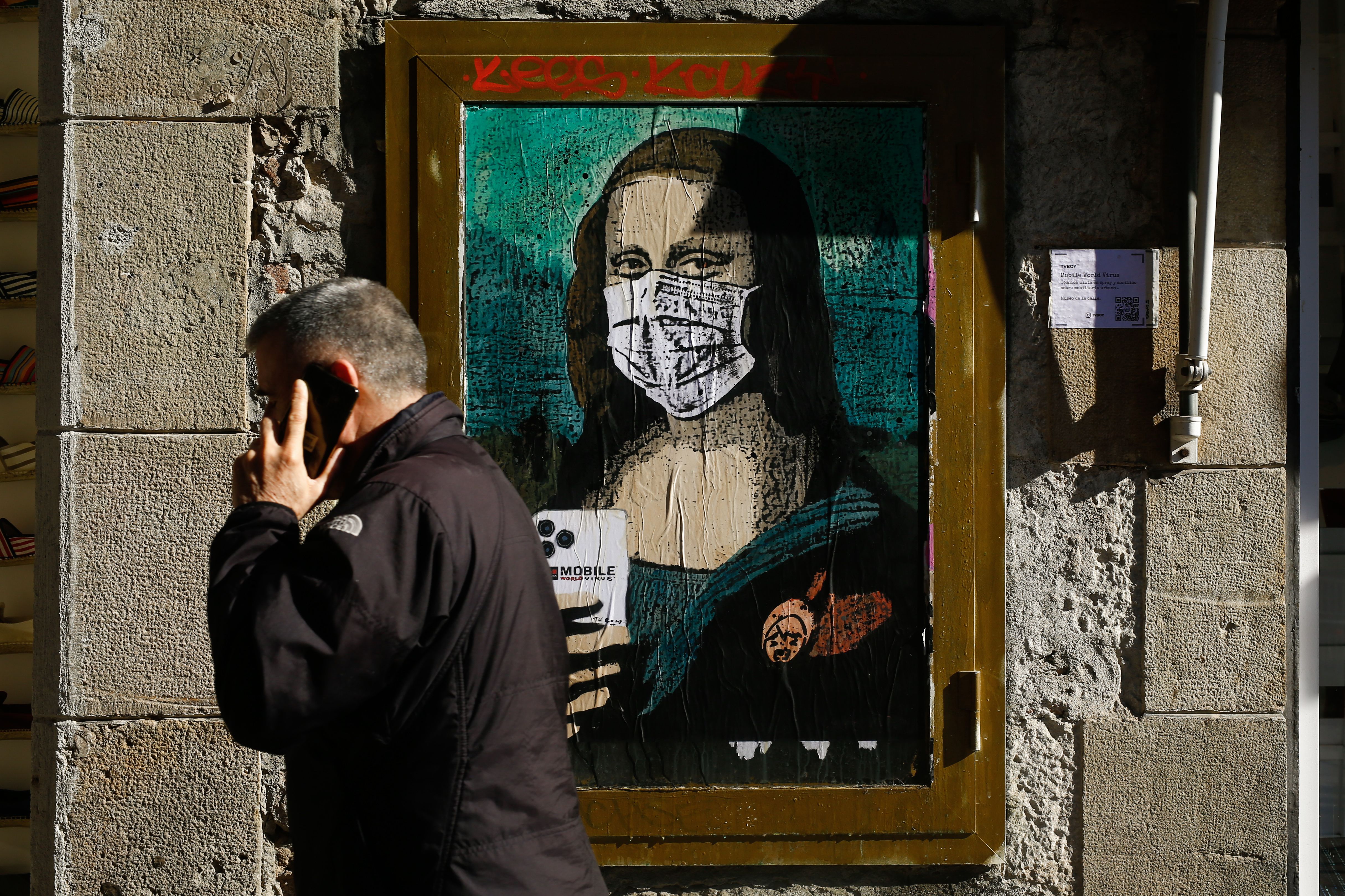 A man holding his mobile phone walks past a poster by Italian urban artist Salvatore Benintende aka "TVBOY" depecting Leonardo da Vinci's Mona Lisa wearing a protective facemask and holding a mobile phone reading "Mobile World Virus" in a street of Barcelona on February 18, 2020, a week after the World Mobile Congress was cancelled due to fears stemming from the coronavirus that sparked an exodus of industry heavyweights. (Photo by PAU BARRENA/AFP via Getty Images)