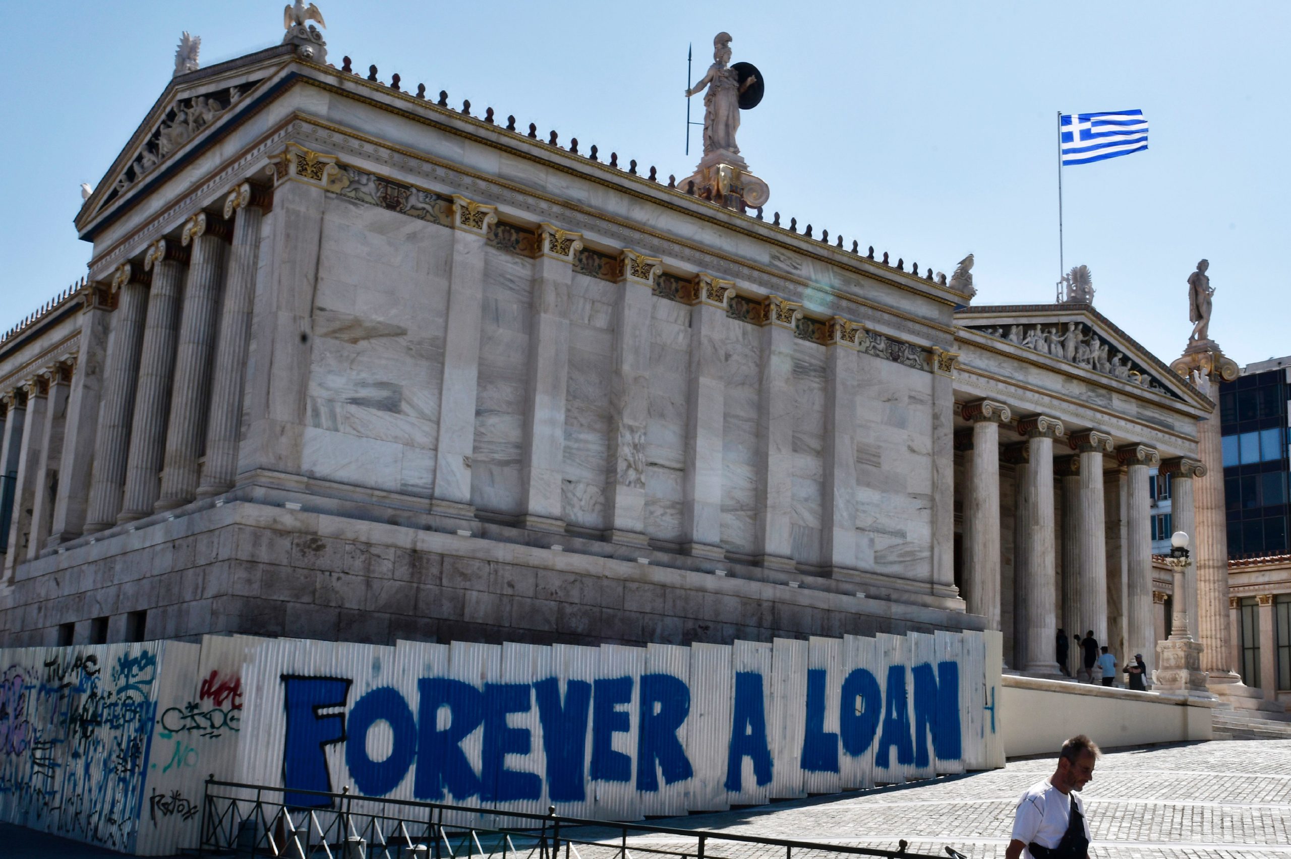 A woman walks past a graffiti refering to the Greek debt and reading "Forever a loan" outside the Academy of Athens building on August 28, 2017. / AFP PHOTO / LOUISA GOULIAMAKI (Photo credit should read LOUISA GOULIAMAKI/AFP via Getty Images)