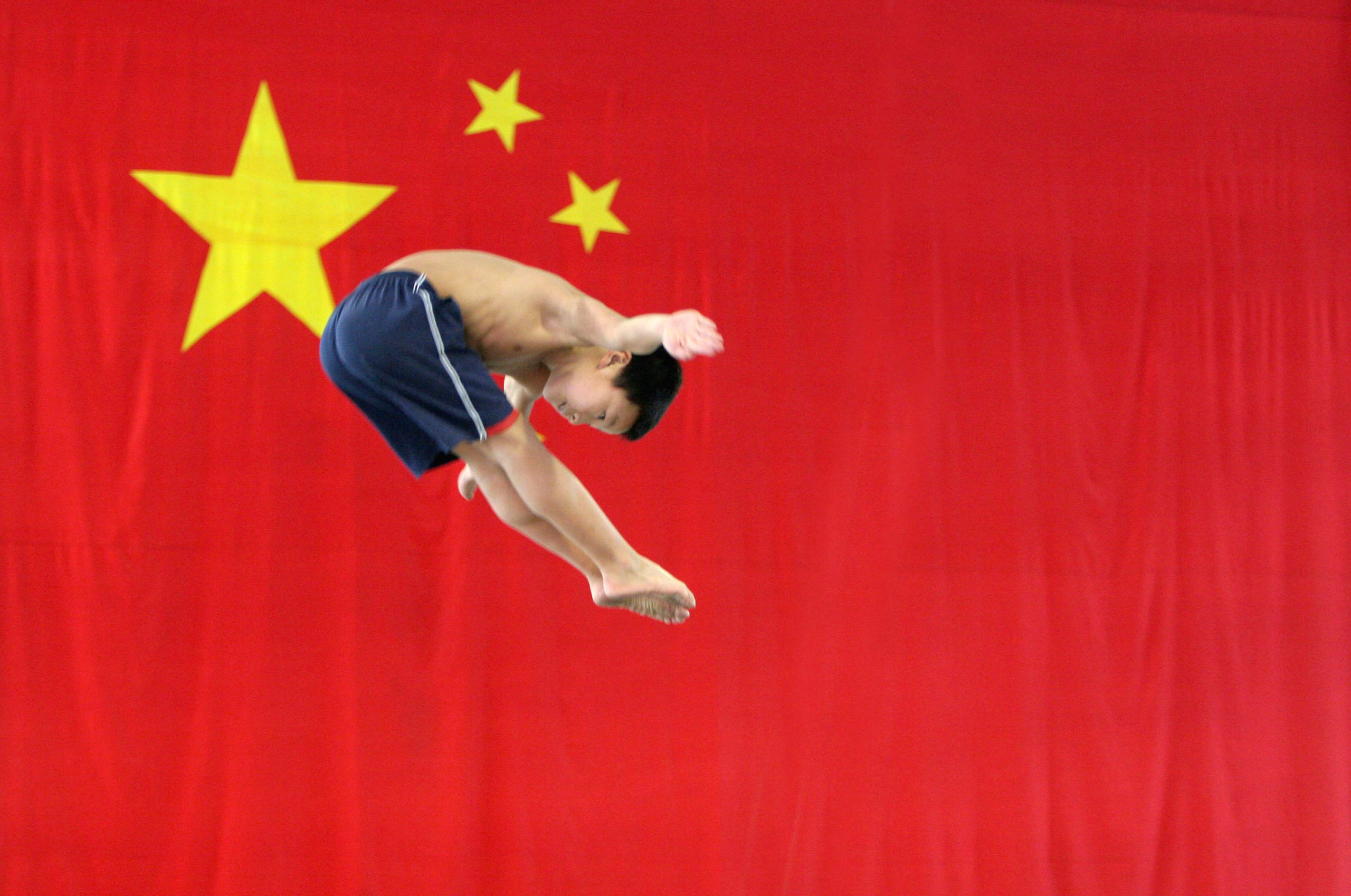 CHENGDU, CHINA - MAY 10: (CHINA OUT) A young boy practises diving movements as he jumps on a trampoline in front of a national flag at a diving school on May 10, 2005 in Chengdu of Sichuan Province, China. There are approximately 3,000 sport schools in China, recruiting thousands of very young children and sending the elites for Olympic training. China has consistently risen in the Olympic medal table, coming second in Athens 2004 and China will host the 2008 Olympic Games. This success has encouraged more parents to send their children to government-funded sport schools, though the training is extremely hard. (Photo by China Photos/Getty Images)