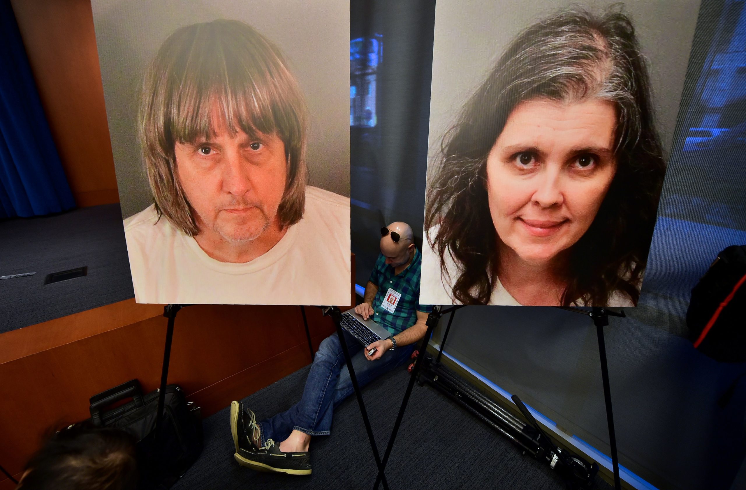 A journalist sits between portraits of David and Louise Turpin awaiting a press briefing in Riverside, California on January 18, 2018. David Allen Turpin, 57, and his wife Louise Anna Turpin, 49 were hit with 12 counts of torture, 12 of false imprisonment, six of child abuse and six of abuse of a dependent adult at a court hearing in the city of Riverside. David Turpin was also charged with committing a lewd act against a child by force or fear or duress, District Attorney Mike Hestrin told a press conference. (FREDERIC J. BROWN/AFP via Getty Images)