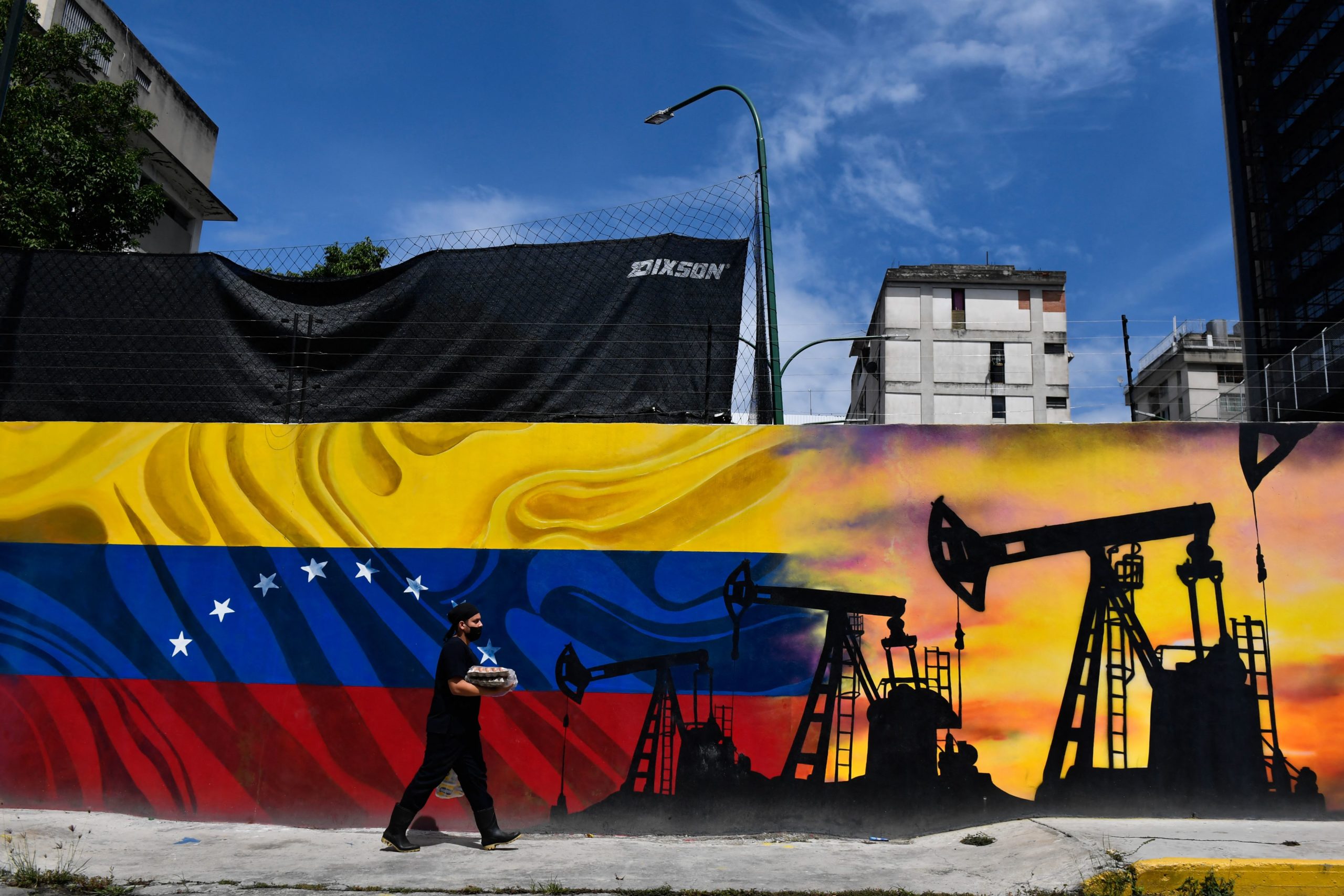 A man wearing a face masks walks past a mural depicting an oil pump and the Venezuelan flag in a street of Caracas, on May 26, 2022. 