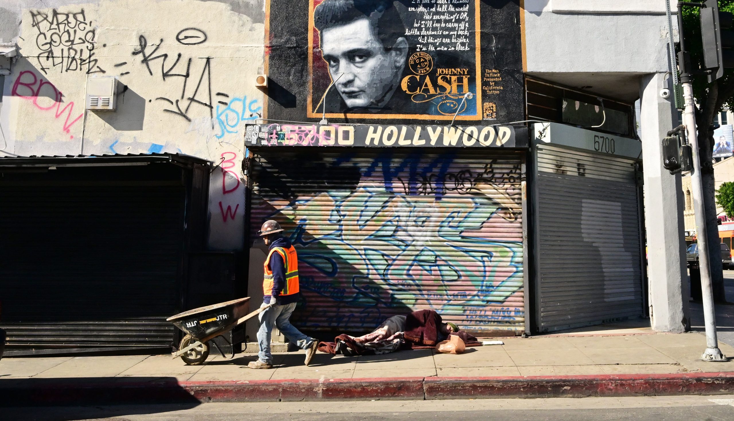 A construction worker pushes his wheelbarrow past a homeless man sleeping on a Hollywood sidewalk on January 20, 2023 in Hollywood, California. - Los Angeles County earlier this month declared a state of emergency over the region's homelessness crisis after decades of increasing numbers of people seen living on the streets.