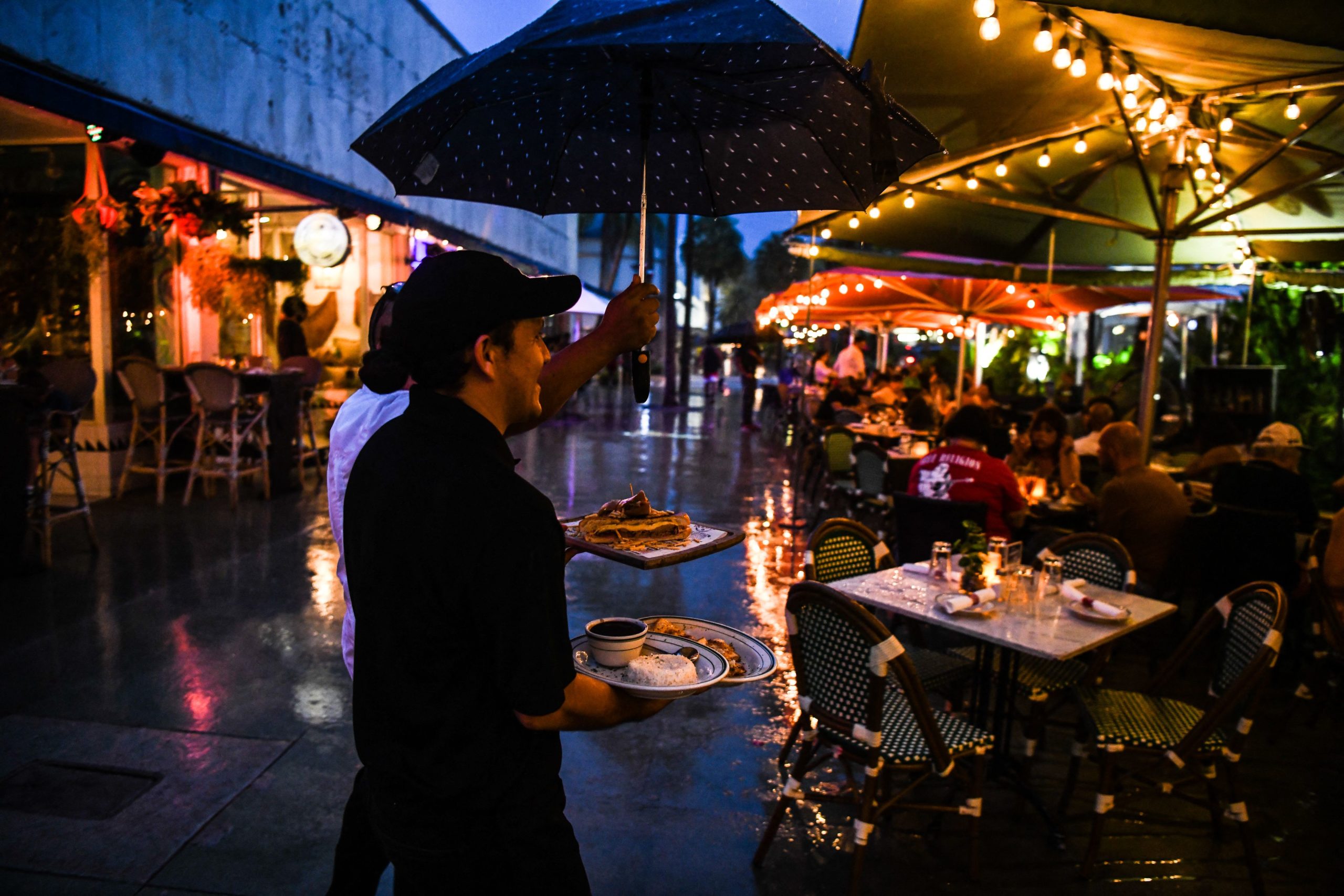 A waiter covers from rain under an umbrella as he delivers food to the table in Miami Beach, on July 27, 2021. (Photo by CHANDAN KHANNA / AFP) (Photo by CHANDAN KHANNA/AFP via Getty Images)