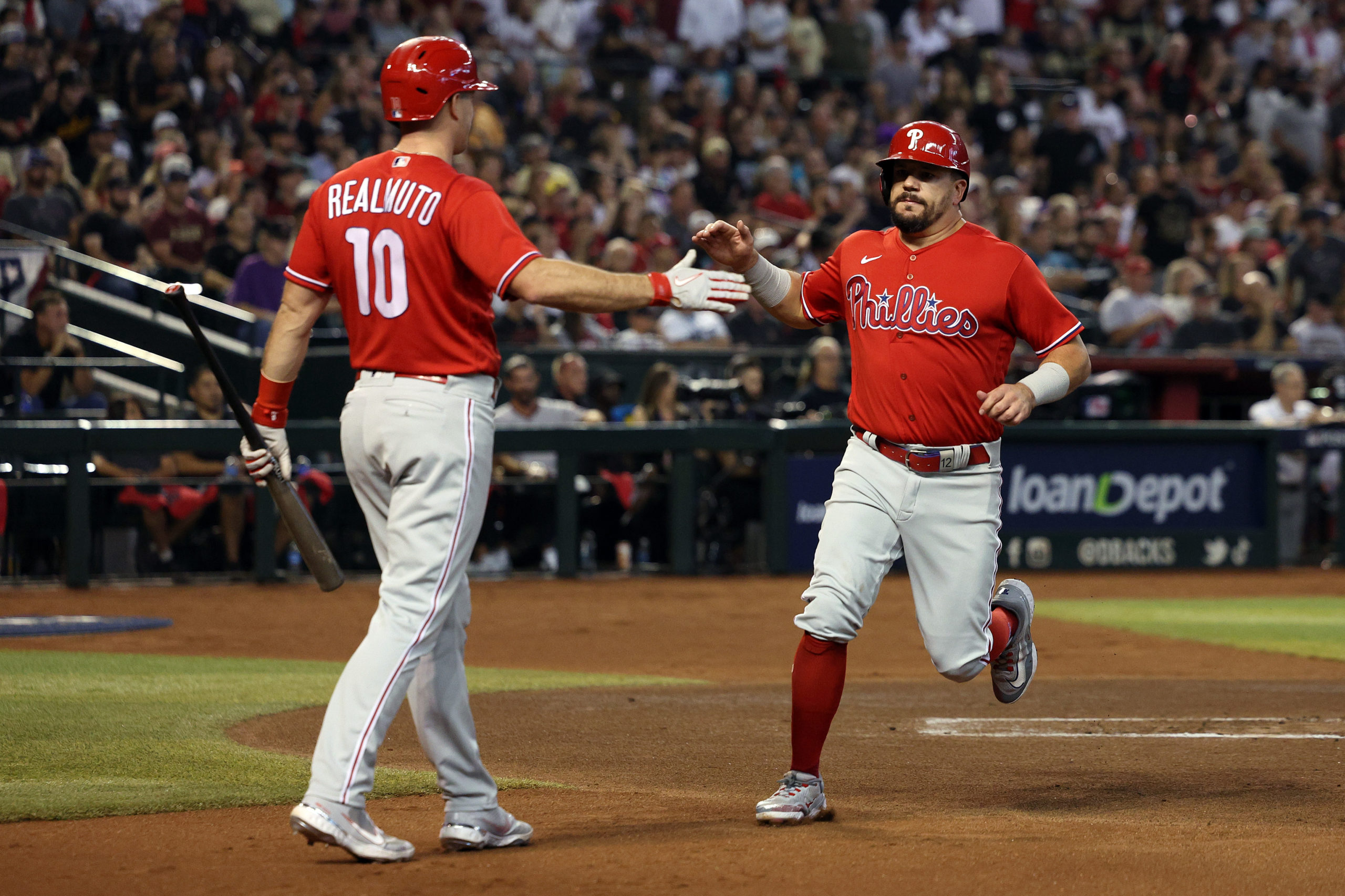 PHOENIX, ARIZONA - OCTOBER 21: Kyle Schwarber #12 of the Philadelphia Phillies celebrates with J.T. Realmuto #10 after scoring a run off of a single by Bryson Stott #5 against the Arizona Diamondbacks during the first inning in Game Five of the National League Championship Series at Chase Field on October 21, 2023 in Phoenix, Arizona. Harry How/Getty Images